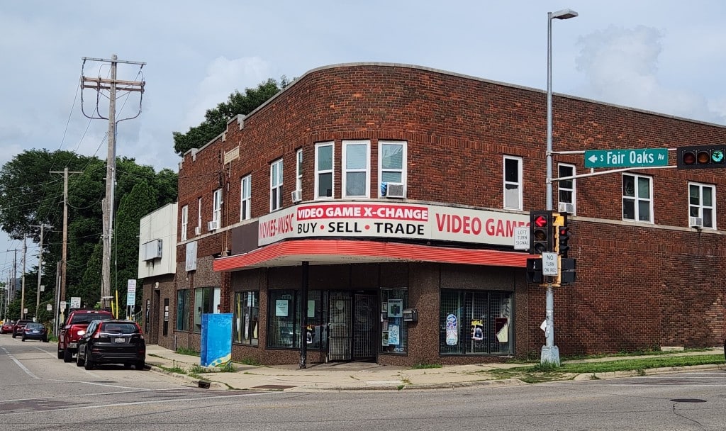 A photo shows the building and storefront of Video Game X-Change at the corner intersection of S Fair Oaks Av and Atwood Av in Madison on a summer afternoon. The large store sign wraps around the side of the building, reading "MOVIES • MUSIC" on the lefthand side and "VIDEO GAMES" on the righthand side in red font. The center of the sign reads "VIDEO GAME X-CHANGE" in white font on a red background. Beneath that are the words "BUY • SELL • TRADE" in black font.