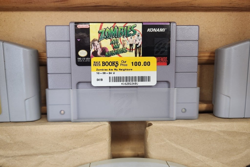 A close-up photo of a grey Super Nintendo video game cartridge sitting on a shelf in a wooden display case. The label on the game shows the title and graphic of "Zombies Ate My Neighbors." A yellow and white Half Price Books price sticker below the label reads "Our Price 100.00" in black font.