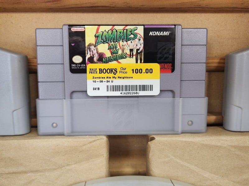 A close-up photo of a grey Super Nintendo video game cartridge sitting on a shelf in a wooden display case. The label on the game shows the title and graphic of "Zombies Ate My Neighbors." A yellow and white Half Price Books price sticker below the label reads "Our Price 100.00" in black font.