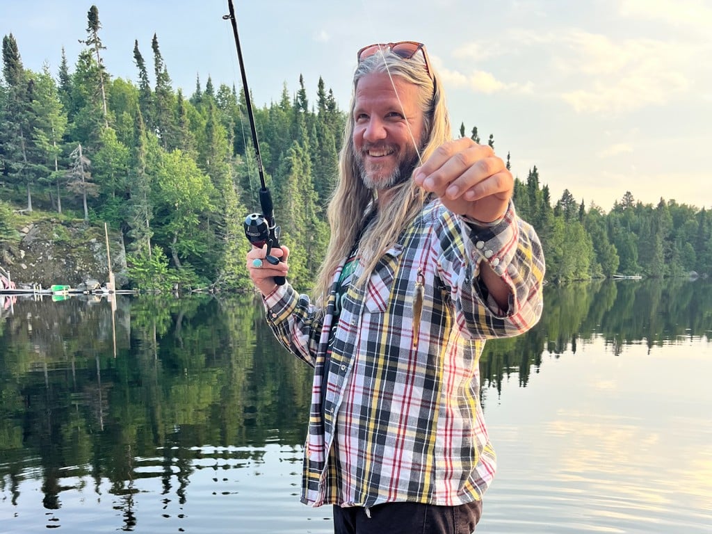 Dan Walkner appears smiling, holding a fishing rod in front of a body of water. He's wearing a plaid shirt and black jeans. His hair is long, he's got a full beard, his sunglasses are perched on top of his head, and he's smiling.