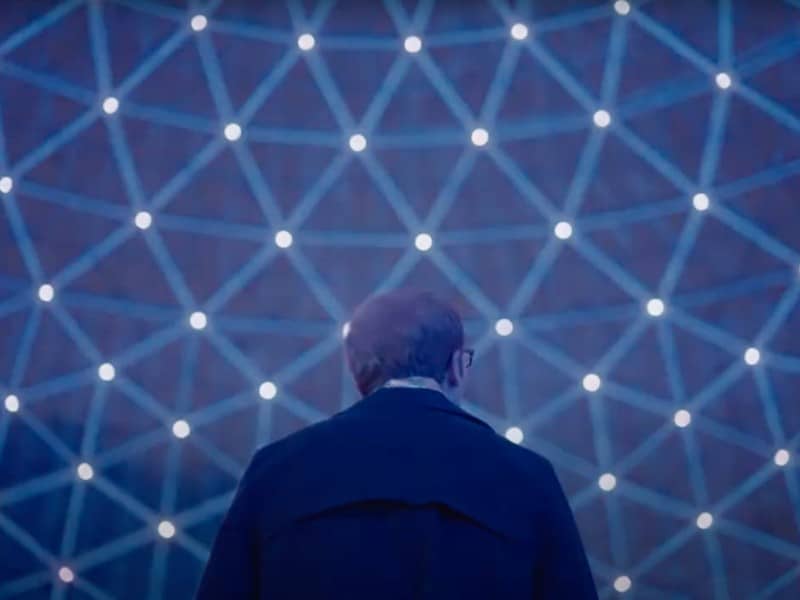 Joe Pera is shown from behind, gazing up at a lights show at Milwaukee's Mitchell Park Domes. There's an interconnected web of lights that illuminate the image in a cool blue. Pera's centered but very low in the frame, amplifying the scope of the Domes. He's wearing a dark, fashionable jacket and his face is not visible.