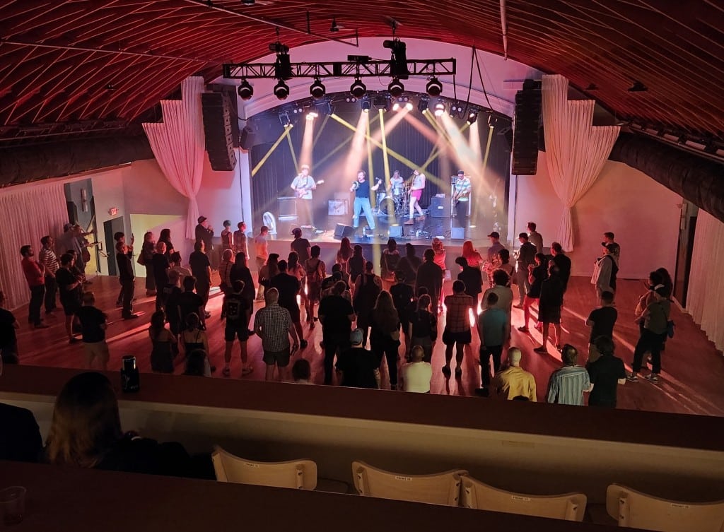 Photo of a performance venue with elaborate stage lighting. Taken at a long shot from a second level, the five-piece band performs on a stage in the background below. A rather large crowd of several dozen people fill out the first floor space in front of the stage.