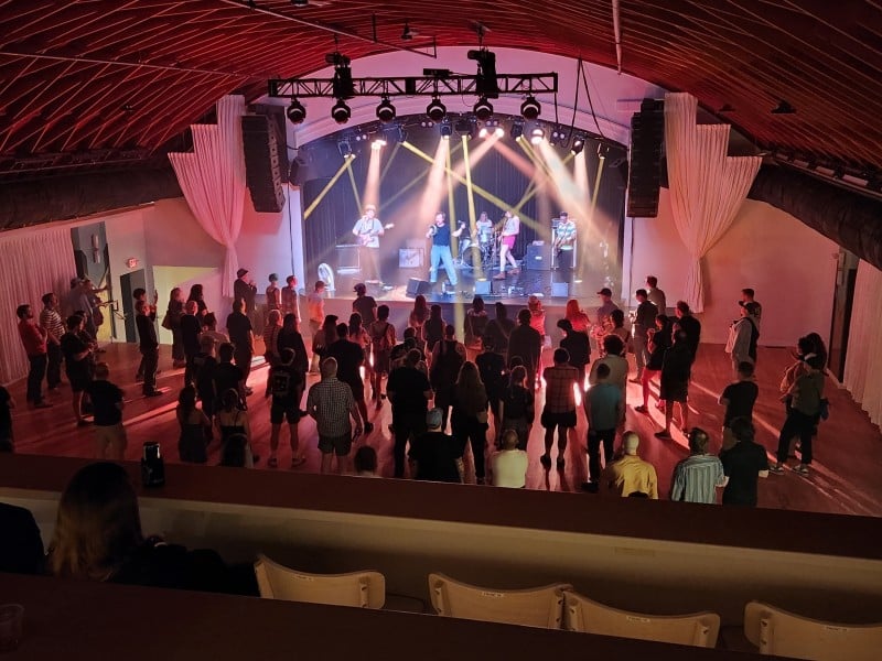 Photo of a performance venue with elaborate stage lighting. Taken at a long shot from a second level, the five-piece band performs on a stage in the background below on the first floor. A rather large crowd of several dozen people fill out the first floor space in front of the stage.