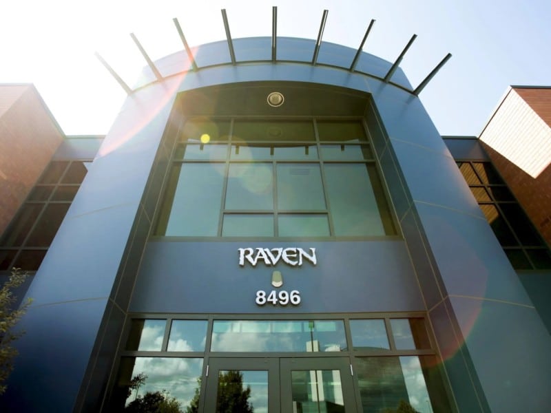 A low-angled photograph of the entrance to a glass building on a sunny day (with lens flare). The company logo of "RAVEN" and number "8496" are mounted above the main doors.