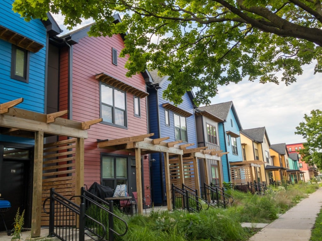 An angled photo shows a row of slim, boxy, similarly designed housing units stretching into the distance. The houses are modest in their appearance, but painted colorfully- including shades of blue, red, yellow, and green at the far end.