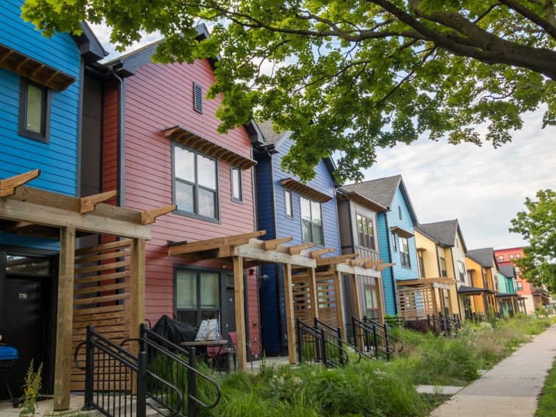 An angled photo shows a row of slim, boxy, similarly designed housing units stretching into the distance. The houses are modest in their appearance, but painted colorfully- including shades of blue, red, yellow, and green at the far end.