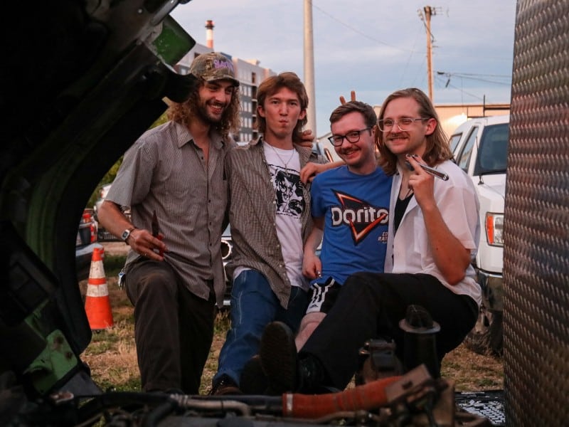 Slick pose with their arms around each other in front of a vehicle being serviced at an auto repair shop. From Left to Right: Calvin Childress, Nate Opperman, Hance Throckmorton, and Morgen Nicodemus. Each band member is holding a small repair tool. They're all smiling, apart from Opperman, whose lips are pursed.