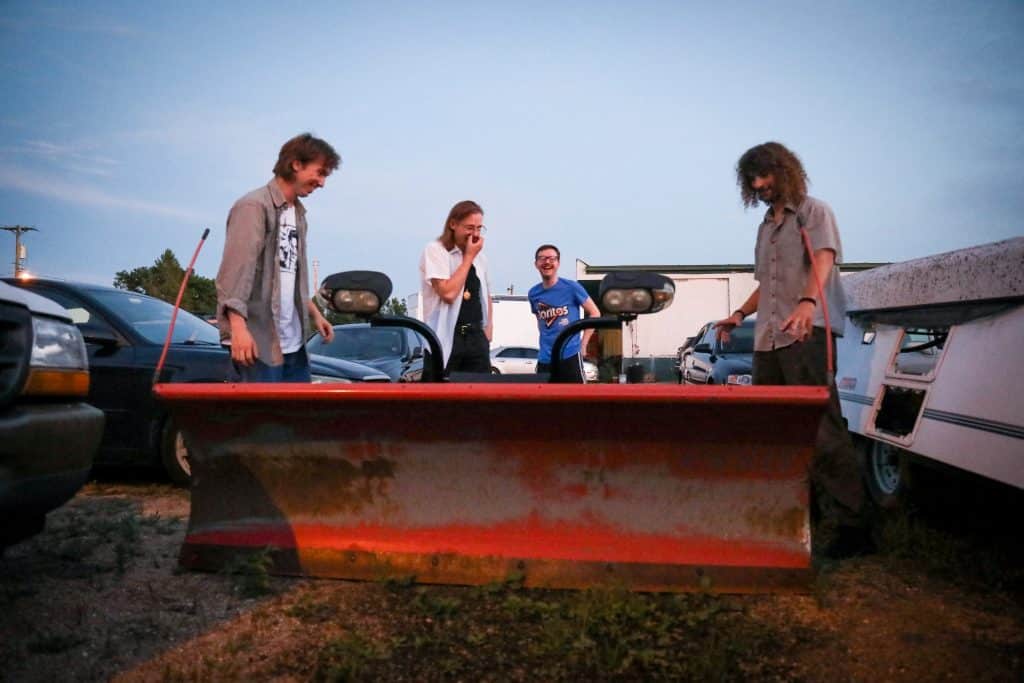 The members of Slick stand around a piece of machinery at an auto repair yard, looking surprised and laughing. From Left to Right: Nate Opperman, Morgen Nicodemus, Hance Throckmorton, and Calvin Childress. 