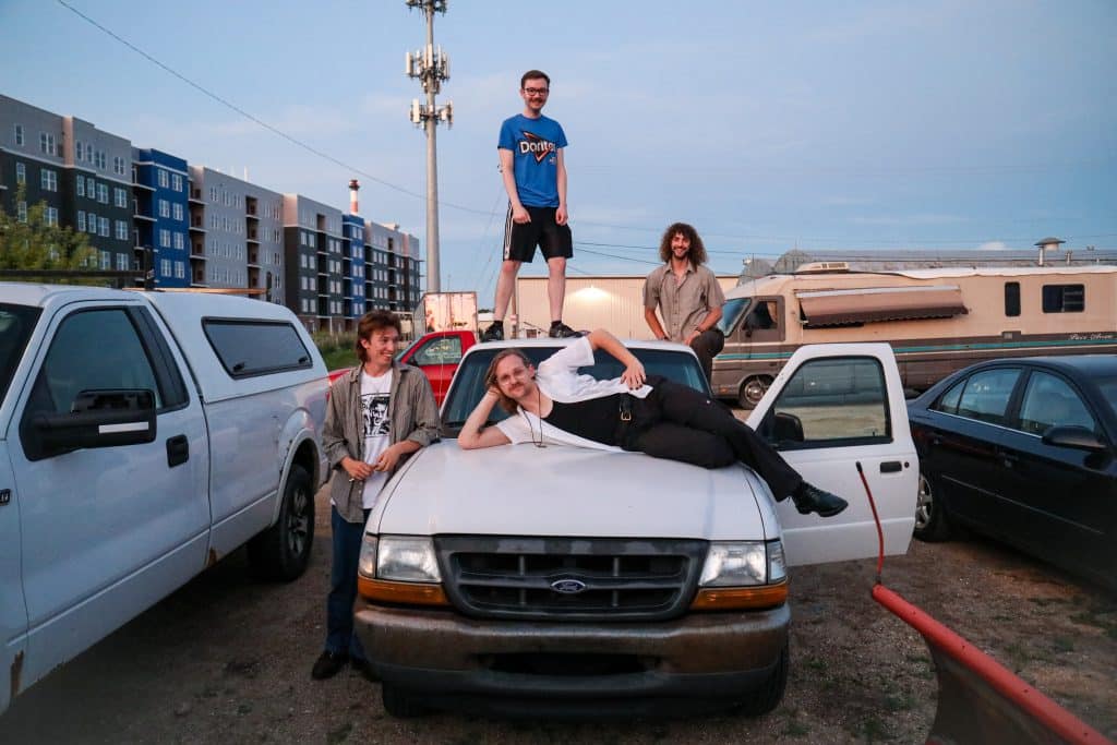 The members of Slick all pose around a white truck. Hance Throckmorton stands on the roof of the truck, wearing glasses and a blue Cool Ranch Doritos shirt. Nate Opperman stands to the left of the truck, leaning into its hood with his elbow. He's wearing an open gray button-up shirt and a white tee with a black graphic. Morgen Nicodemus is laying horizontally across the truck's hood, sporting a necklace, an open white button-up, a black shirt, and black pants. Calvin Childress is standing on the truck's bed, and his leg is only partially visible. He's wearing a beige button-up and dark pants. All members are smiling. 