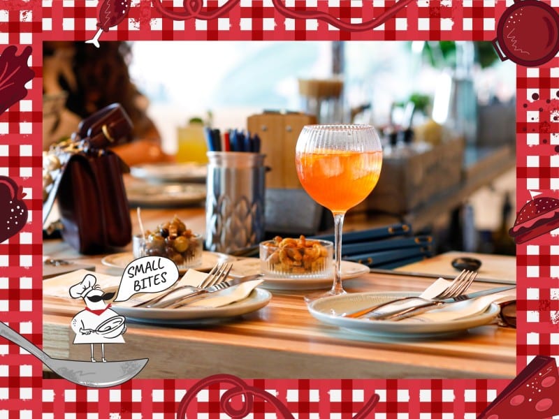 A professional photo of a table arrangement at an upscale restaurant at a slight angle. Several small plates with silverware and appetizers sit atop a thick wooden countertop. A wine glass with an orange-looking beverage sits in between the plates. Behind them, in the background and to the left, are various boxes and containers that are out of focus. Bordering this photo is a red and white checkered tablecloth illustration. In the lower left corner of this illustrated frame is a small chef with an oversized mustache standing on a spoon. The chef's speech bubble reads "Small Bites."