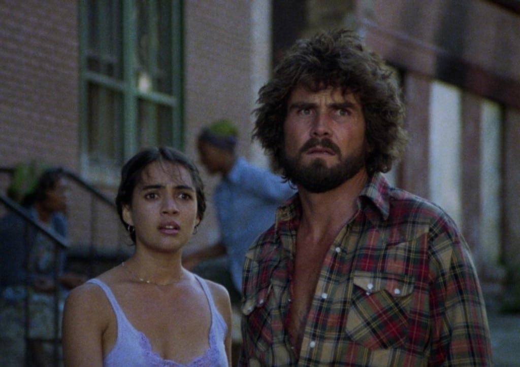 A still image from the 1980 film "Night Of The Juggler" shows a man and a woman standing at a medium close-up in an area of the South Bronx, New York City. They stare off into the distance and look worn out in the waning daylight. The woman on the left wears a thin, pale blue tank top. The bearded man on the right wears a heavier red and gold plaid shirt that is partly unbuttoned.