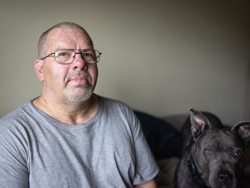 A medium close-up photo of a middle-aged man sitting on a couch in his living room with his dog to his right. He wears a grey t-shirt and looks up and at the photographer with a neutral expression.