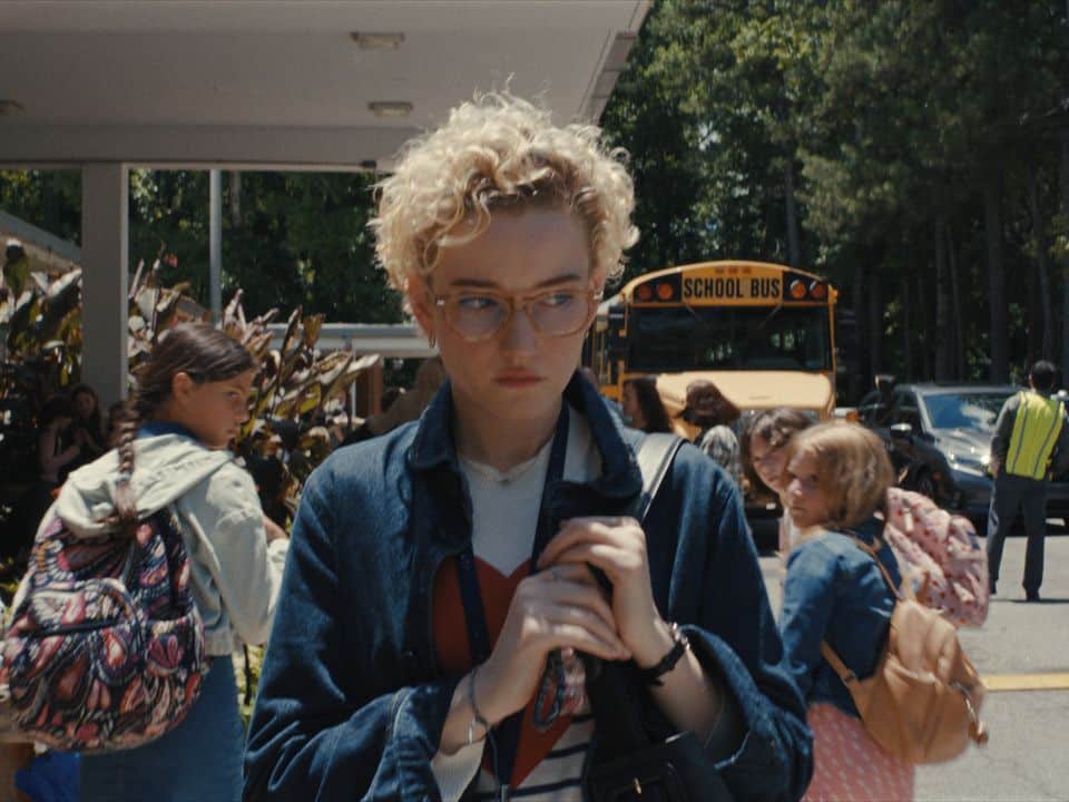 A still from the film "Weapons" shows a medium shot of character Justine Gandy walking across a school parking lot with an apprehensive expression. Justine has short and curly blonde hair, and she wears a light denim jacket and holds onto the strap of a black bag thrown over her left shoulder. Several kids behind her turn and stare at her with suspicion.