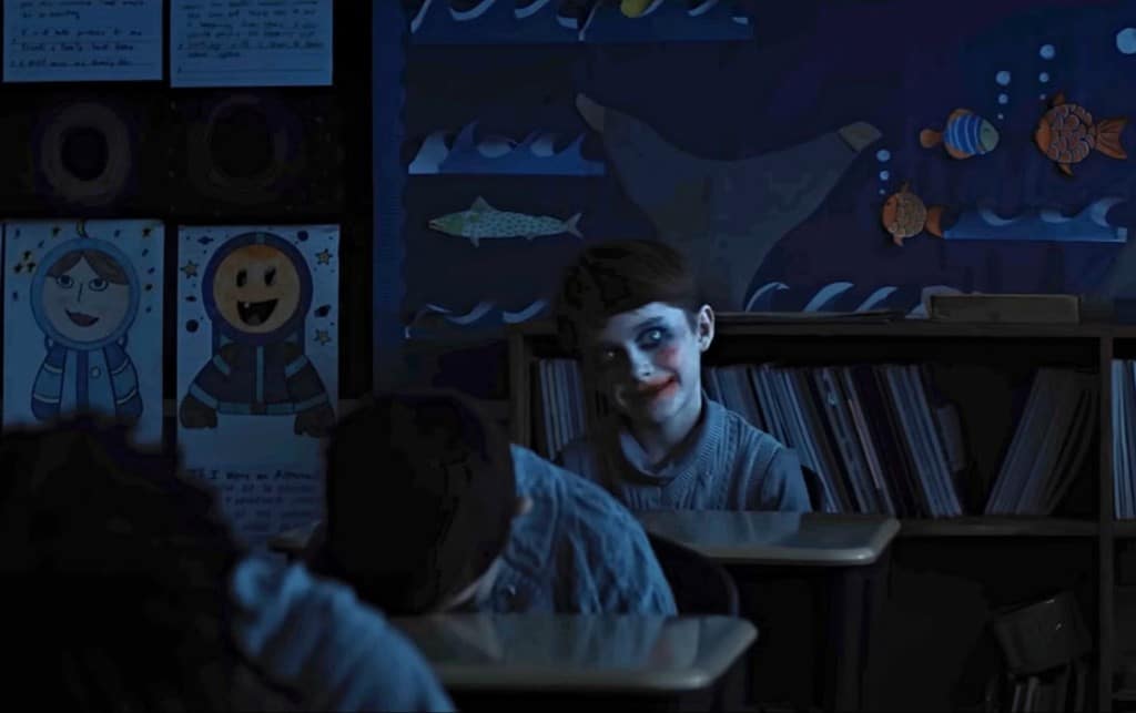 A dimly lit still image from the film "Weapons" shows an elementary-school classroom with an aquatic mural and student art posted on the back wall. Two students in the foreground have their heads down on their desks as if they're sleeping, while the student sitting in the back row is wide awake with a deranged, wide-eyed smile. He wears clown-like face paint and stares forward.