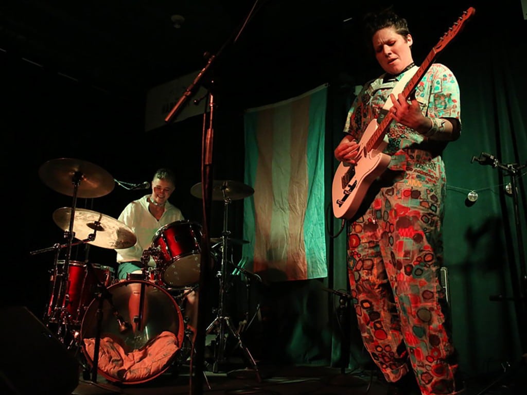 Damsel Trash performs at Harmony Bar & Grill in 2024. Emily Mills is playing a red-shell drum kit and smiling to the image's left. To their right, Meghan Rose is playing a white Telecaster and scrunching up her face in concentration. In the center of the image, behind the duo, a trans flag hangs on the venue's wall. Mills is wearing a chunky necklace and a white zip-up jacket. Rose is wearing a colorfully patterned jumpsuit. Mills, Rose, and the flag are illuminated, the rest of the image is nearly black, with only some stray stage equipment visible.