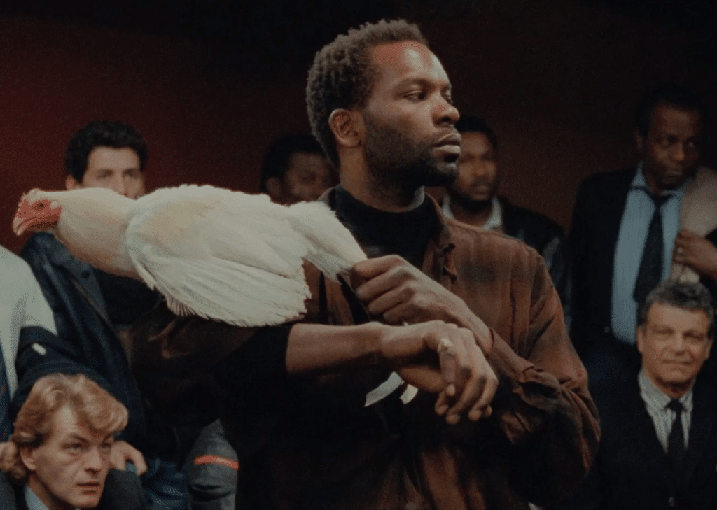 A screenshot of "No Fear, No Die" shows the Black character, Jocelyn, standing before a diverse audience of cockfighting spectators surrounding him. Jocelyn holds a white rooster, Toni, over his shoulder in a dominant, flaunting pose.