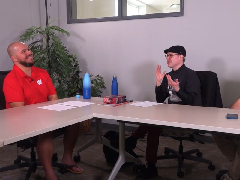 At a medium shot, two people sit in a simple conference room at two elongated desks arranged in a triangular-like formation. The two people are turned towards one another, smiling jovially. Paper notes, steel water bottles, and a phone are placed on the tables in front of or to the side of the people.