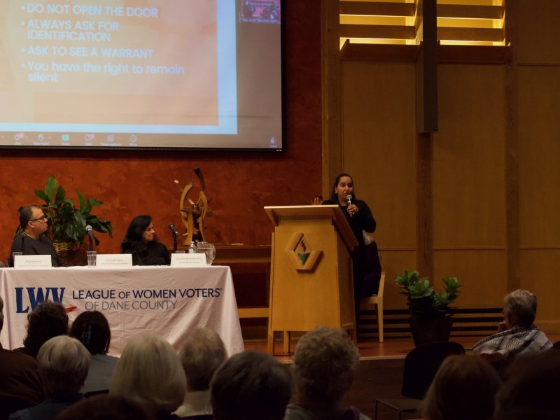Photograph at a medium-long shot of a large conference room where an audience is seated in the foreground and facing the stage. In the background, on the stage, two panelists are seated at a table with a white banner thrown over it that reads "LWV League of Women Voters of Dane County." The two panelists turn towards the female speaker standing at a wooden lectern. She faces the audience and holds a microphone. On the far wall behind them is a projection screen that displays a presentation's bullet points.