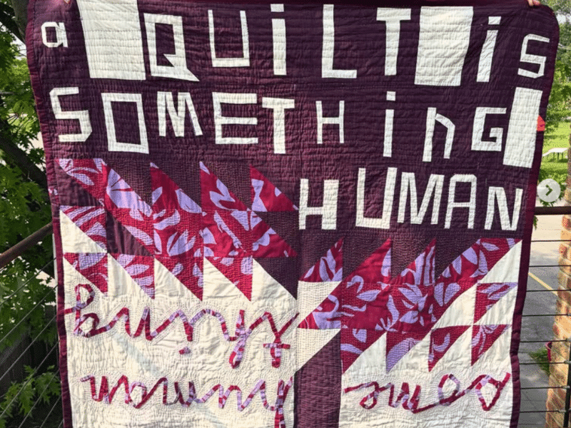 A maroon and white quilt with floral triangles reads "a QuiLT is SOMETHinG HUMAN" in custom printed lettering. Upside down, the quilt also reads "some human thing" in a continuous cursive font. The quilt is displayed vertically on an outdoor balcony.
