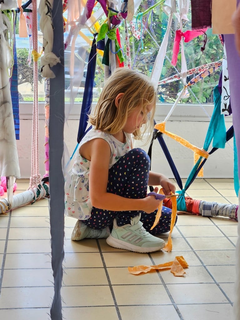 A young girl kneels on the floor of a sunlit room. She cuts a yellow fabric with scissors. Other strips of fabric are woven together and hang above and around her from PVC piping, which is constructed into a boxy frame-like shape.