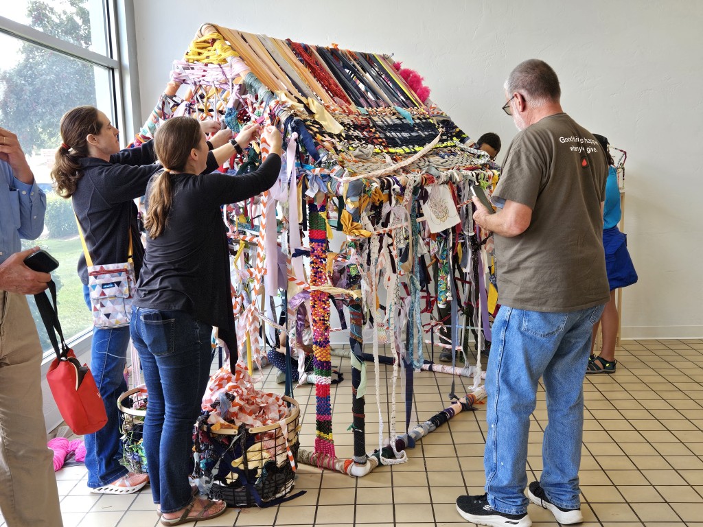 Several people of various ages, all dressed in casual clothes, stand in a sunlit room. They are all tying or weaving strips of fabric to decorate PVC pipes that are constructed into a frame in a house shape.