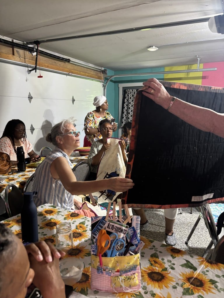 In a fluorescent-lit room with a white garage door in the background, several women are quilting and examining materials. The women near the center of the frame looks to the right to examine a quilt that someone off-screen is holding up.