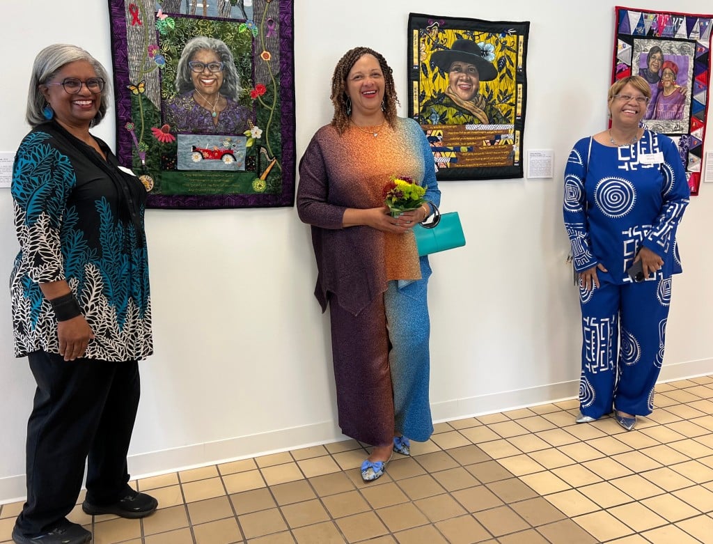 Three women in colorful, artistically designed garments stand near a gallery wall with their elaborate quilts on display. The quilts include beautiful floral designs and dignified self-portraits. The three women face the camera and smile.