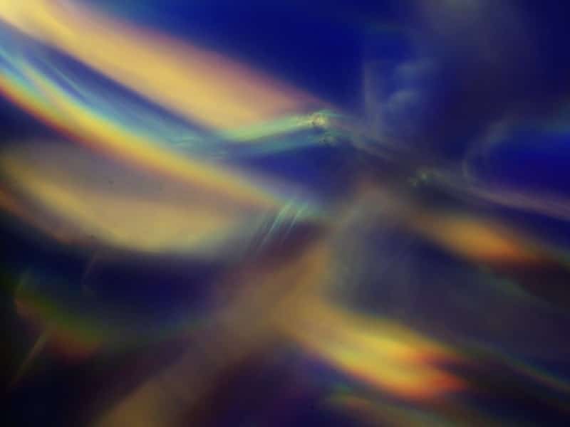 An abstract, prismatic still from Blake Barit's short film "Journey To Sunrise." It features a blurry blend of blues, golds, greens, and black to form a vague, interpretative image of insect wings in motion or sunstreaking in a dark environment.