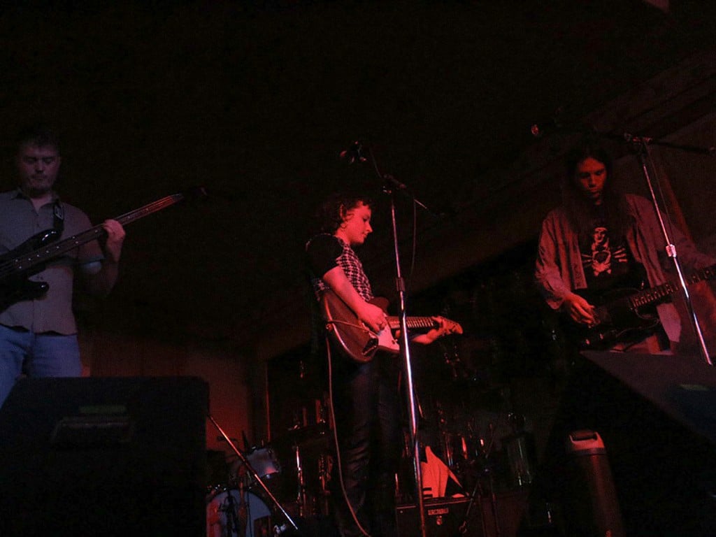 Jane Hobson performs live at the Crystal Corner Bar in 2023. The photograph is faded. In it, Hobson is centerstage, playing guitar. To her left is a bassist, to her right a guitarist. The band's drummer is not clearly visible, hidden in part by a large stage monitor on the image's bottom left corner.