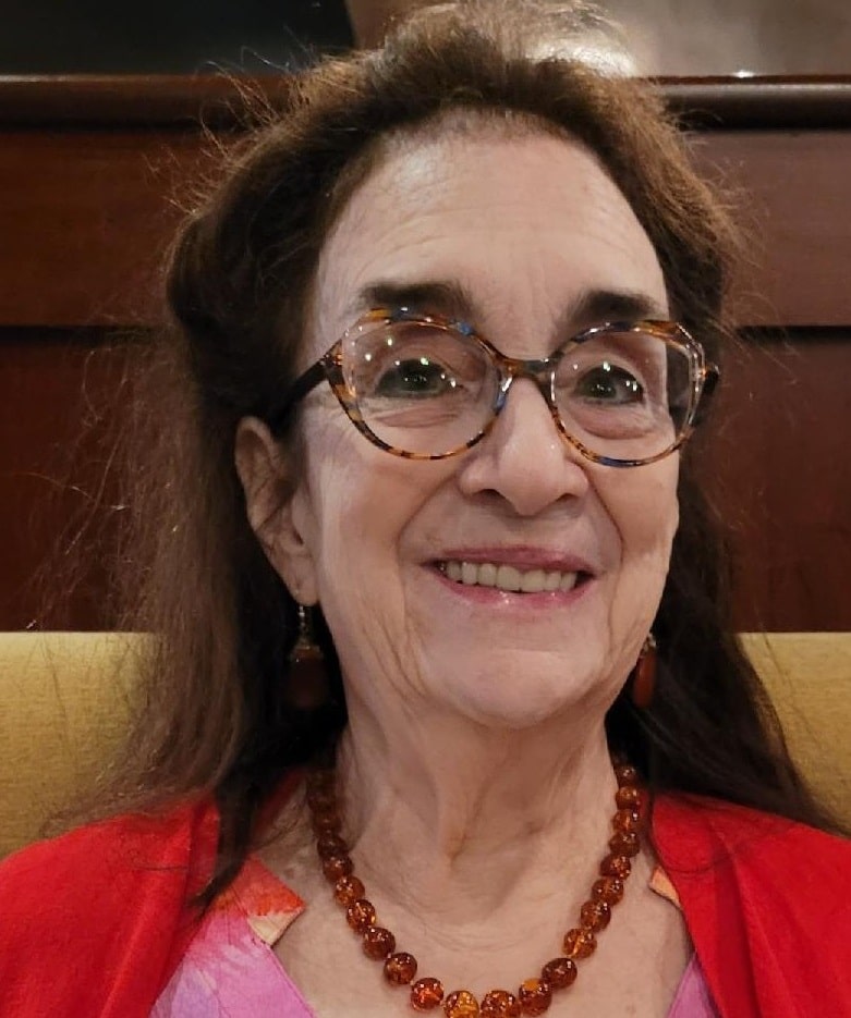 Close-up headshot of playwright and director Jan Levine Thal, who appears to be sitting on a mustard-colored chair or couch. She wears a redish brown beaded necklace, bright red cardigan, and slightly oversized tortoise-shell patterned glasses. She smiles while looking forward at the camera.