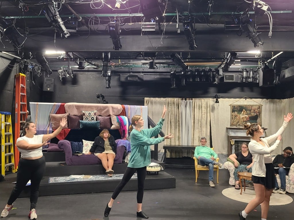A medium-long shot photo of a rehearsal stage contains seven actors in everyday clothing. Four of them are seated on chairs and couches behind three dancers in the foreground who all strike similar poses. The dancers are turned towards the right and gesture upward with their arms. Hanging above the actors, the lighting and tech equipment can be seen.