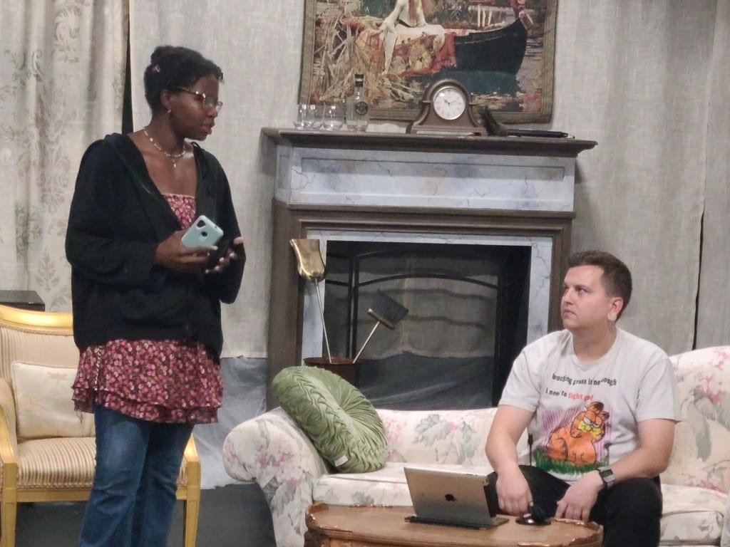 A medium-shot photo captures two actors in everyday clothing rehearsing a scene on a furnished stage that contains a couch, chair, fireplace, and a mantle. The woman, holding a smart phone in her right hand, stands at the left and looks down and to the right at a man sitting on a couch. He returns her gaze.