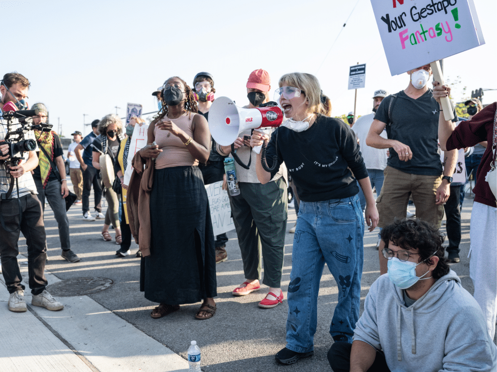 A crowd of protestors gather in the street. They include Illinois Congressional candidate Kat Abughazaleh (at the center-right), who shouts into a white and red megaphone. Other protestors carry signs while wearing face masks and eye protection.