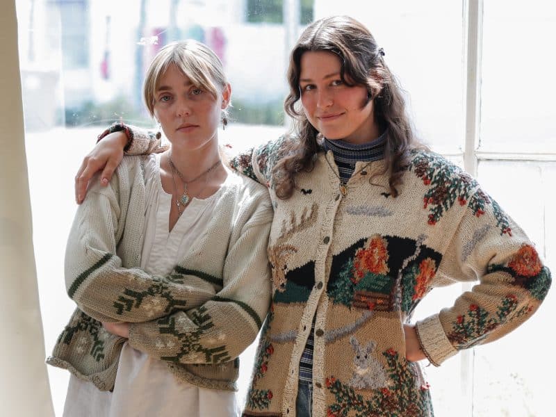 Kate Ruland (Left) and Emma O'Shea (Right) of The Spine Stealers are shown in their Madison studio. Both are wearing cream-colored sweaters with wintry designs. O'Shea has one arm around Ruland's shoulders and another on her hip, and is smiling at the camera. Ruland wears a stoic expression, and has her arms crossed around her chest. Both are wearing pendant necklaces. The sun shines through the window behind them, casting a subtle light on their hair and shoulders.
