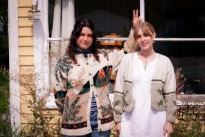 The Spine Stealers' Emma O'Shea and Kate Ruland pose in front of their Madison studio. They're both wearing wintry sweaters, smiling at the camera. O'Shea to the left, Ruland to the right. O'Shea is doing bunny ears above Ruland's head. The image is soft, slightly faded, and has a subtle grain effect.