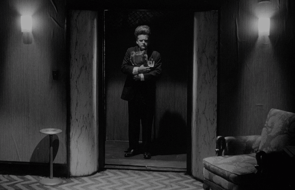 A black-and-white still from the film "Eraserhead" shows an apprehensive man with staticky hair. He's dressed in a dark suit and stands in an opened elevator. The floor in front of him has a pair of hallway lights on the wall and a chevron floor pattern that the director David Lynch came to be known for.
