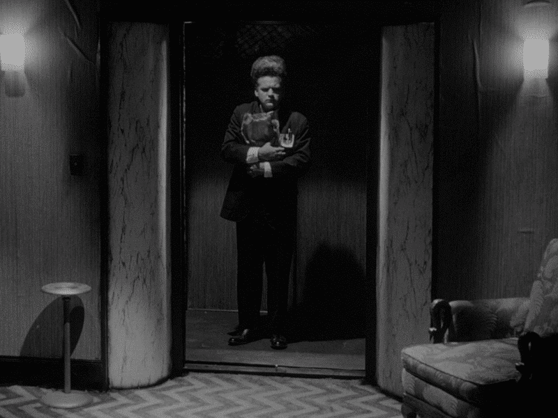 A black-and-white still from the film "Eraserhead" shows an apprehensive man with staticky hair. He's dressed in a dark suit and stands in an opened elevator. The floor in front of him has a pair of hallway lights on the wall and a chevron floor pattern that the director David Lynch came to be known for.
