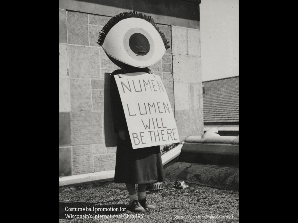 A historic black-and-white photograph shows a female body wearing a giant eye that covers their head and a posterboard sign that reads "Numen lumen will be there."