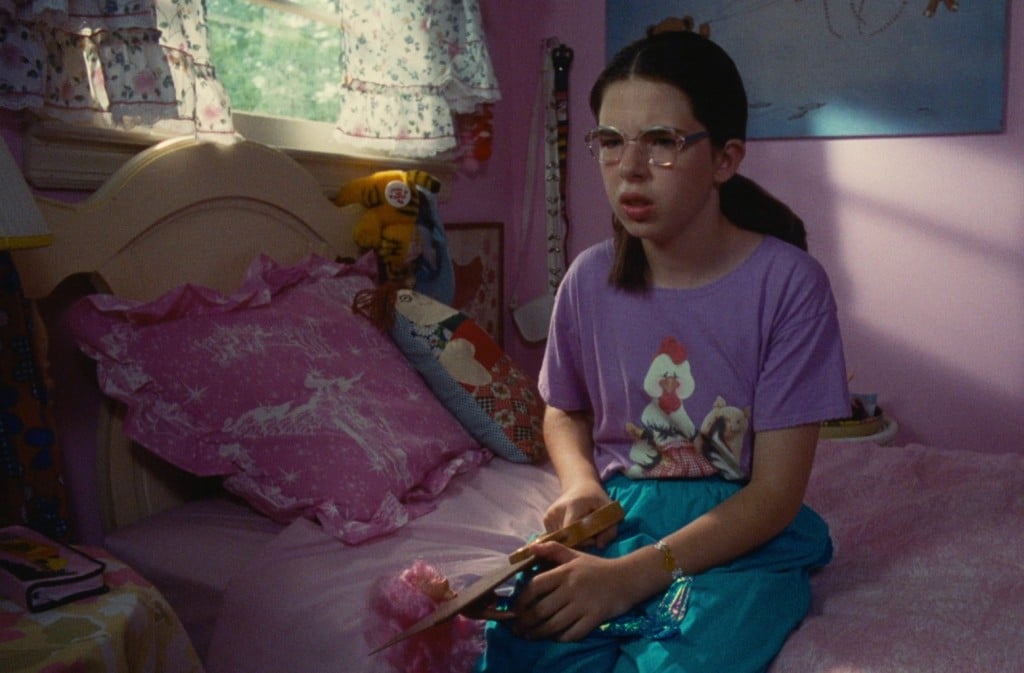 A still from the film "Welcome To The Dollhouse" shows a bespectacled pre-teen girl sitting on her pink bedspread in a shared bedroom. She wears a kitschy pastel-purple animal T-shirt with turquoise-colored pants, and holds a handsaw up to the neck of her sister's Barbie doll on her lap. She stares forward with a blank, yet distressed look.