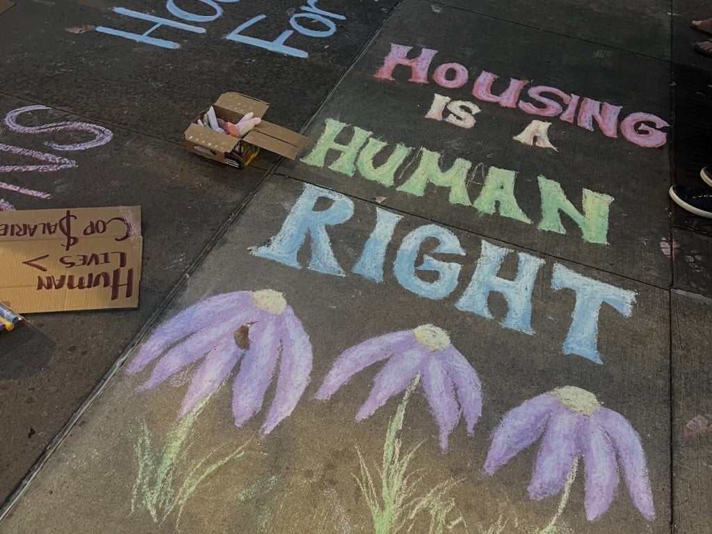 An angled photos shows a grey-black sidewalk that contains an empowering, activist message drawn in various red, yellow, green, and blue chalk. In large capital letters, it reads "HOUSING IS A HUMAN RIGHT." Three purple perennial flowers are drawn below it. To the left of the message is a box of chalk and an upside-down cardboard sign.