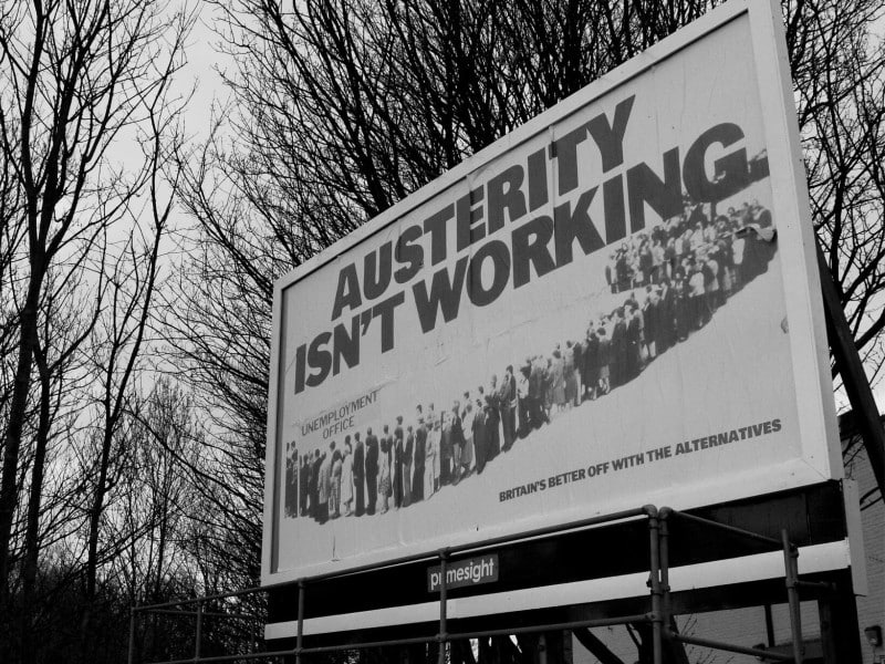 A black-and-white photo of a large outdoor billboard tilted at a left angle. In large black font, it reads "AUSTERITY ISN'T WORKING." Below that, a winding crowd of people wait at the unemployment office. In the bottom right corner, smaller black font reads "Britain's better off with the alternatives."