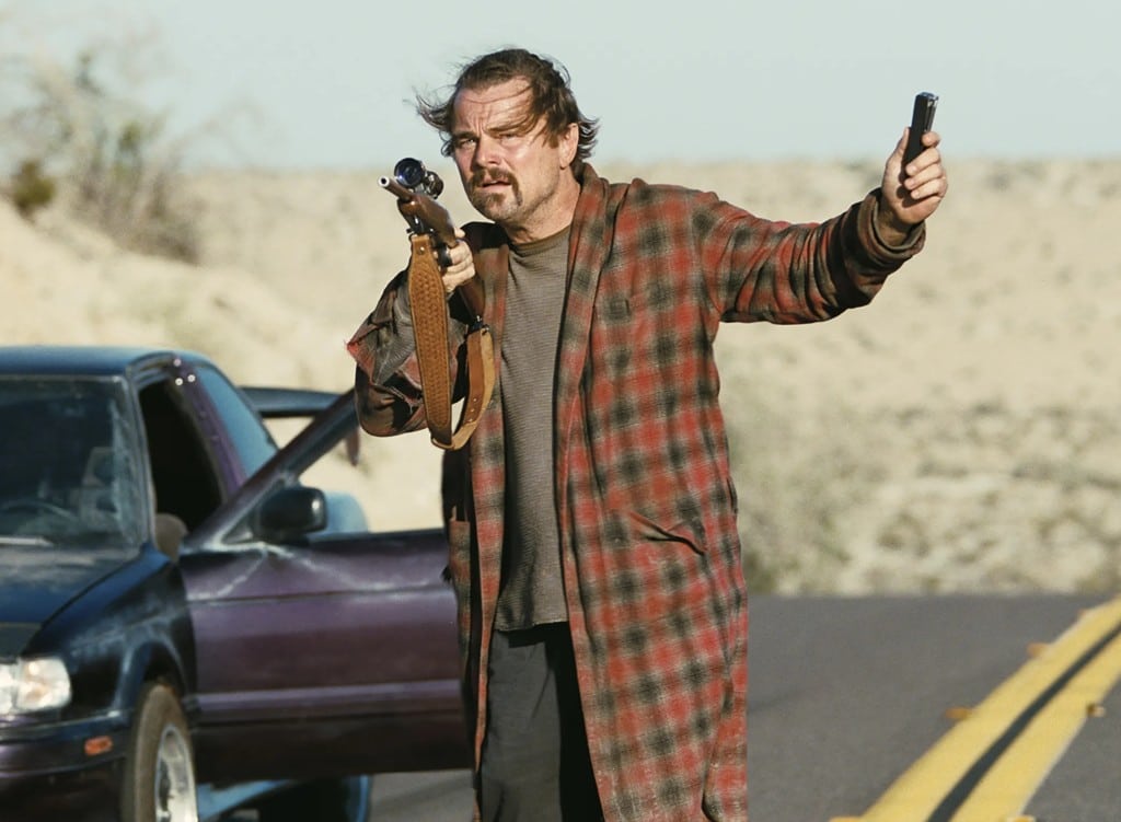 A cropped still from the film "One Battle After Another" shows a disheveled middle-aged man standing on an open desert road. He wears a plaid-patterned bathrobe over a plain grey shirt and black pants. He holds a rifle up with his right arm and holds out a small black device in his left. The man peers into the distance with a concerned expression. A sports car that he's driven is parked in the middle of the road with the driver's side door open.
