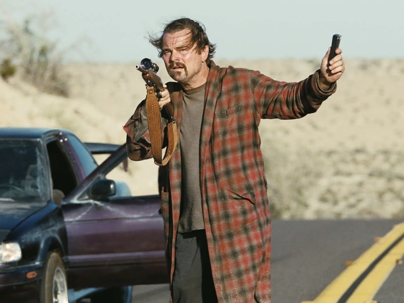 A cropped still from the film "One Battle After Another" shows a disheveled middle-aged man standing on an open desert road. He wears a plaid-patterned bathrobe over a plain grey shirt and black pants. He holds a rifle up with his right arm and holds out a small black device in his left. The man peers into the distance with a concerned expression. A sports car that he's driven is parked in the middle of the road with the driver's side door open.
