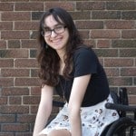 Photo of a young white woman with long dark hair, bangs, and glasses seated in a wheelchair.