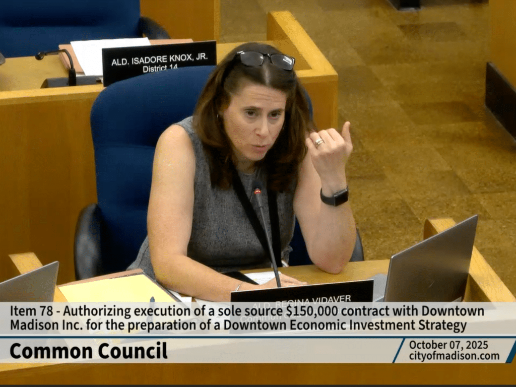 A white woman with shoulder-length brown hair sits at desk. She raises her left hand upward in a motion and leans forward slightly to speak into a microphone. A chairon at the bottom of the image explains that she is at a Madison Common Council meeting on October 7, 2025.
