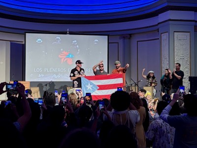 A trio of men stand centerstage holding up a Puerto Rican flag and pointing out to a dimly lit audience clapping and holding up phones to record.