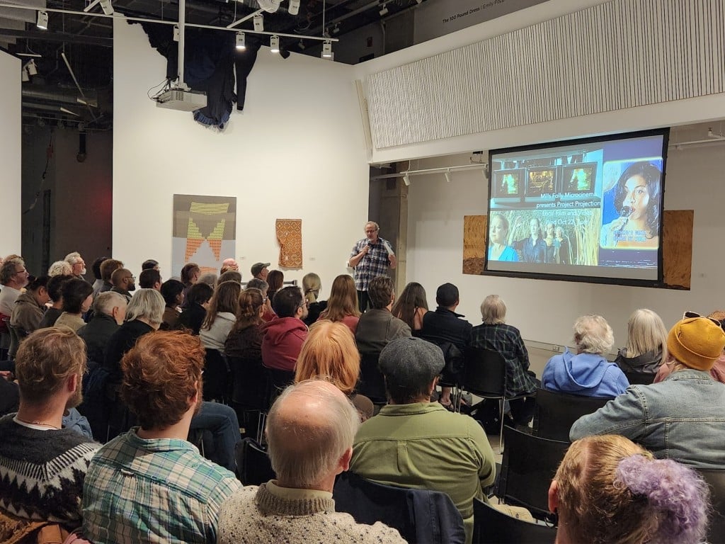 An angled photo at an art gallery shows a large crowd of seated people, who are all looking forward and listening to a male speaker with a microphone at the front corner of the room (who is centered in this photo). To the speaker's right is a projection screen that displays a promotional collage of stills from short films in the forthcoming program.