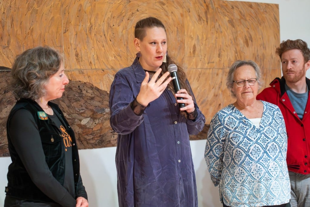 A professional photo at a medium shot shows a woman, filmmaker and choreographer Erica Pinigis, in a long purple blouse standing in an art gallery. She holds a microphone with her left hand and extends forward to gesture with her right hand, while looking out to the right at an unseen audience. Three other filmmakers stand beside her with neutral expressions, turned slightly towards her to listen.
