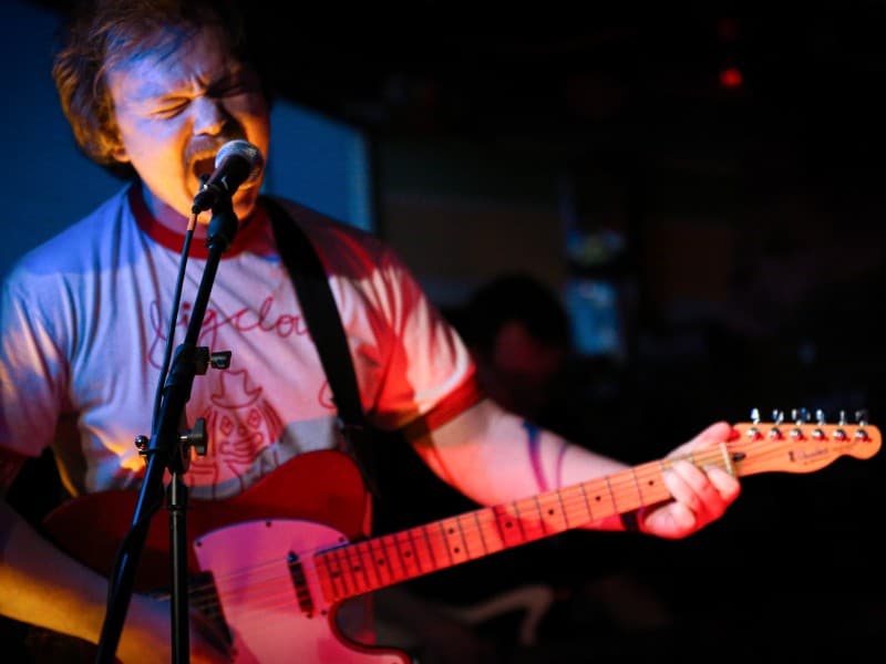Tyler Fassnacht performs live at GBUFO Museum Gift Shop and Records in 2025 as part of Heather The Jerk. His face is scrunched as he plays guitar and screams into the microphone. He's somewhat out of focus, while the microphone stand's arm lock is in focus in front of him. He's wearing a white band shirt with a red illustration and red collar. Behind him is nearly all black. A ghostly blue cascades over Fassnacht's person (and red Fender Telecaster) as he sings.