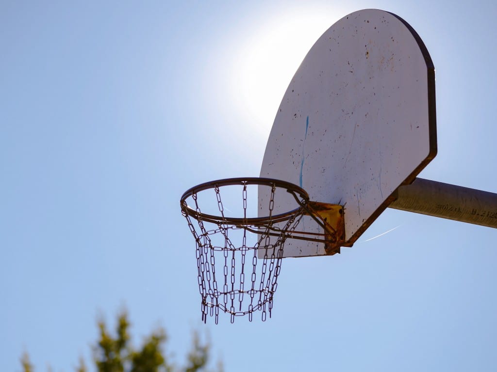 A close-up of a basketball hoop at Windom Way Park in Madison, WI. The hoop takes up the center of the image. The sky is blue and the sun is positioned behind the backboard, providing a glow that helps illuminate the image. The rim is rusty and the net is metal chainlink. The backboard's an aged off-white with fading paint splatter. In the bottom left corner of the image, some treetops are visible, but out of focus.